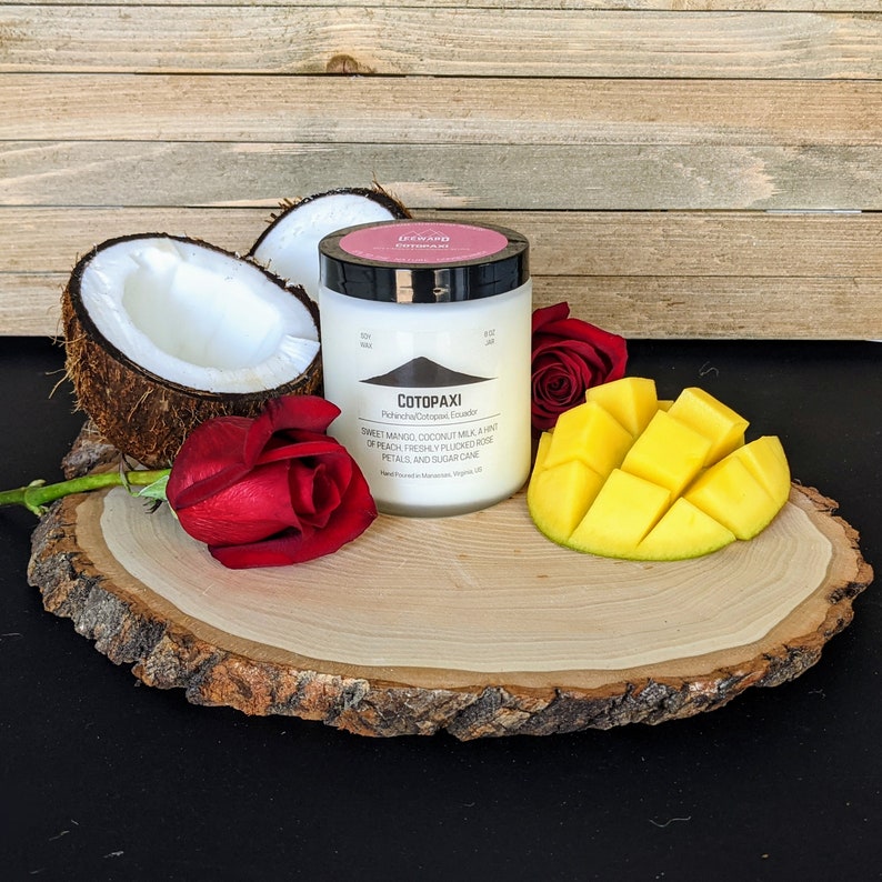 Może przedstawiać: Biały słoik świecy Cotopaxi z czarną pokrywką, otoczony poł&oacute;wką kokosa, czerwonymi r&oacute;żami i pokrojoną mango na drewnianej desce. Etykieta zawiera składniki i nazwę marki.