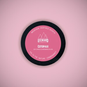 Może przedstawiać: Okrągła pokrywka na świecę z czarną obw&oacute;dką i r&oacute;żowym środkiem. Na środku znajduje się napis "COTOPAXI" i "MOUNTAIN INSPIRED SCENTS". Świeca jest na bazie soi i ręcznie robiona w USA. Marka to Leeward Candle Co.