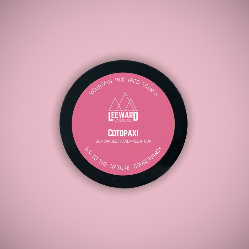 Może przedstawiać: Okrągła pokrywka na świecę z czarną obw&oacute;dką i r&oacute;żowym środkiem. Na środku znajduje się napis "COTOPAXI" i "MOUNTAIN INSPIRED SCENTS". Świeca jest na bazie soi i ręcznie robiona w USA. Marka to Leeward Candle Co.