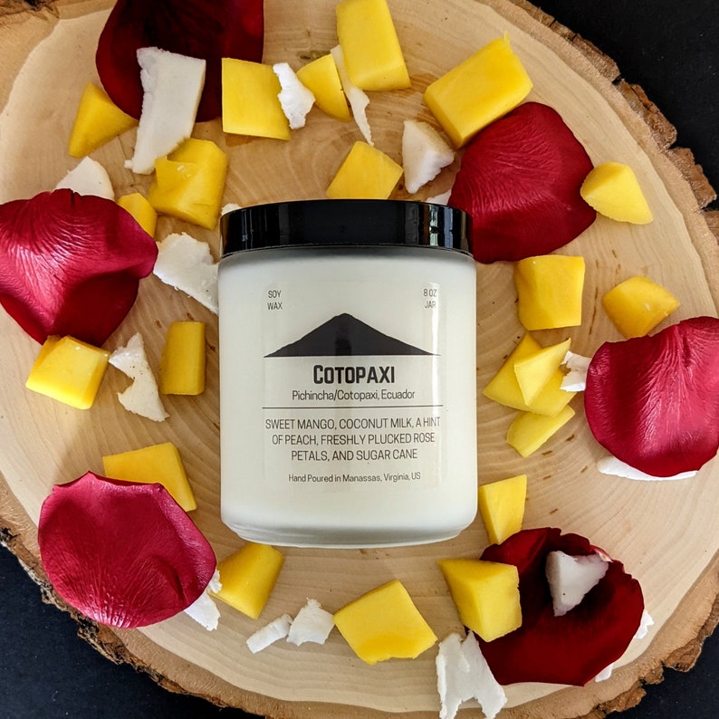 Może przedstawiać: Świeca z wosku sojowego w przezroczystym słoiku z czarną pokrywką, oznaczona jako "COTOPAXI". Świeca otoczona jest czerwonymi płatkami r&oacute;ż, ż&oacute;łtymi kawałkami mango i białymi kawałkami kokosa na drewnianej powierzchni. Etykieta wymienia zapach słodkiego mango, mleka kokosowego, brzoskwini, płatk&oacute;w r&oacute;ż i trzciny cukrowej.
