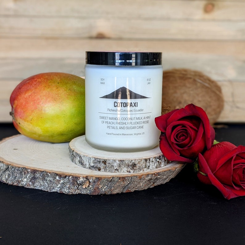 Może przedstawiać: Świeca z wosku sojowego w szklanym słoiku z czarną pokrywką, z etykietą "COTOPAXI" z nutami zapachowymi. Mango, kokos i dwie czerwone r&oacute;że leżą obok świecy na drewnianym plastrze.