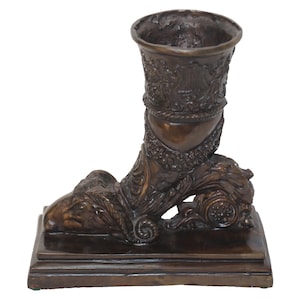 May include: A bronze sculpture of a stylized boot with a decorative vase on top. The boot is detailed with intricate carvings and rests on a square base.