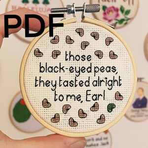 May include: A close-up of a round embroidery hoop with a white fabric background. The fabric has brown and green embroidered hearts and the text "those black-eyed peas, they tasted alright to me, Earl".