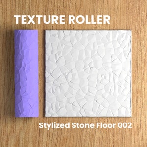 Puede incluir: Un rodillo de textura morado con un patrón de suelo de piedra estilizado. El rodillo se utiliza para crear una superficie texturizada en paredes o suelos. El texto "TEXTURE ROLLER" está por encima del rodillo y el texto "Stylized Stone Floor 002" está por debajo del rodillo.