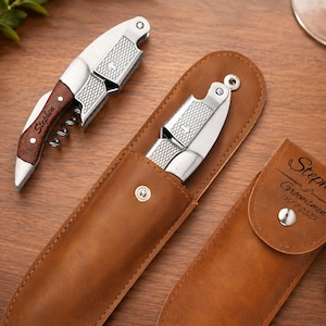 Puede incluir: Dos sacacorchos de plata y madera con espiral y cortacápsulas, uno de ellos dentro de una funda de cuero marrón. El mango de madera tiene grabado el nombre Stephen. La funda de cuero tiene un broche metálico y el texto Stephen Groomsmen 06.07.2025.