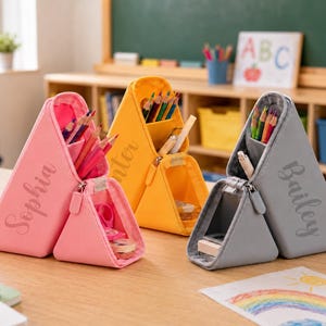 May include: Three triangular pencil cases in pink, yellow, and gray, each featuring a personalized name in script font. The cases have a main compartment for pencils and a smaller front zippered section for accessories, standing upright on a desk.