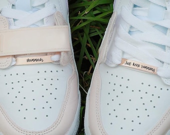May include: Pair of white and light purple sneakers with cream-colored accents, sitting on grass. Each shoe has a cream-colored strap with text. One says "Hannah" and the other says "Just keep running".