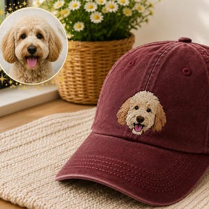 May include: A maroon baseball cap featuring an embroidered patch of a light brown curly-haired dog with its tongue out. The cap has a structured crown, a curved brim, and stitched eyelets on the top panels, showcasing a detailed canine portrait.