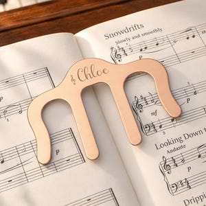 May include: A rose gold metal music book page holder with a curved, three-pronged design. The centre of the holder features the engraved name Chloe next to a small treble clef symbol. It is resting on an open book of printed musical sheet music.
