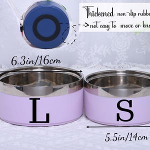 K&ouml;nnte beinhalten: Zwei Edelstahl-Lunchboxen mit lilafarbenen Deckeln. Die gr&ouml;&szlig;ere Box ist mit "L" gekennzeichnet und hat einen Durchmesser von 16 cm und eine H&ouml;he von 7,5 cm. Die kleinere Box ist mit "S" gekennzeichnet und hat einen Durchmesser von 14 cm und eine H&ouml;he von 7 cm.