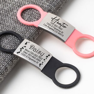 K&ouml;nnte beinhalten: Zwei personalisierte Haustier-ID-Tags mit schwarzen und rosa Silikonringen. Das schwarze Schild tr&auml;gt den Namen "Daisy" und die Telefonnummern von Mama und Papa sowie die Adresse "1333 S Figueroa St". Das rosa Schild tr&auml;gt den Namen "Hunter" und die Adresse "1333 S Figueroa St, Los Angeles, CA 90015" sowie die Telefonnummer "720-352-7902".