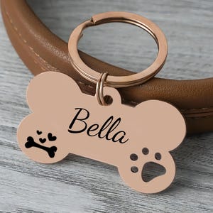 May include: A rose gold dog tag in the shape of a bone, with the name "Bella" in black script. The tag features a paw print and a bone with hearts. Attached to a rose gold keyring.