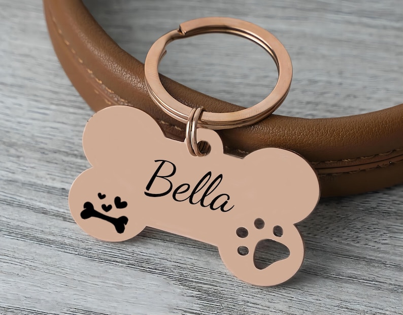 May include: A rose gold dog tag in the shape of a bone, with the name "Bella" in black script. The tag features a paw print and a bone with hearts. Attached to a rose gold keyring.