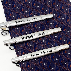 Op de afbeelding: Drie zilveren stropdasclips met gegraveerde tekst. De bovenste clip luidt "Jason 8.25.2024", de middelste clip luidt "best man | Jason" en de onderste clip luidt "Aaron Chagall".