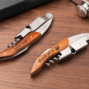 May include: Two stainless steel corkscrews with wooden handles. One handle is engraved with the text 2014 . 7.15 and the other with the text Grand-père and a hand gesture icon. Both tools feature a folding mechanism and a metal spiral worm.