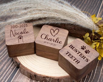 May include: Three square wooden boxes with engraved text and designs. One box reads "Nicole" with a paw print, another says "Charlie" with a heart, and the third says "Buster's Teeth" with paw prints. The boxes are on a wooden slice.