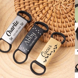 Puede incluir: Tres accesorios de etiquetas personalizadas para mascotas con lazos elásticos negros. Las etiquetas son plateadas, negras y doradas, cada una con el nombre y número de teléfono de la mascota. La etiqueta plateada dice "Charlie" con detalles de mariposas. La etiqueta negra dice "Abby".