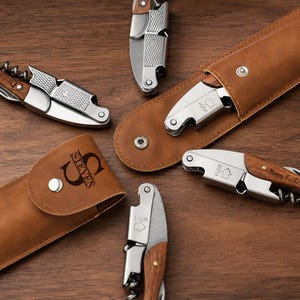May include: Several stainless steel corkscrews with wooden handles, some featuring engraved text like "STEVEN" and "Happy Fathers Day". Two corkscrews are stored in tan leather pouches with silver snap closures, while others are displayed open.