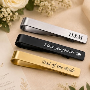 May include: Three metal tie clips in silver, black, and gold tones. The silver clip is engraved with H&M, the black clip features the text I love you forever with a sun icon, and the gold clip is engraved with the text Dad of the Bride.