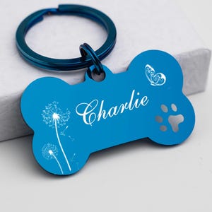 Puede incluir: Chapa para perro de hueso azul con un llavero plateado. La chapa tiene un dise&ntilde;o de diente de le&oacute;n blanco, una mariposa, una huella de pata y el nombre "Charlie" grabado.