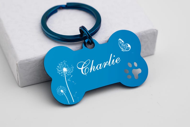 Puede incluir: Chapa para perro de hueso azul con un llavero plateado. La chapa tiene un dise&ntilde;o de diente de le&oacute;n blanco, una mariposa, una huella de pata y el nombre "Charlie" grabado.