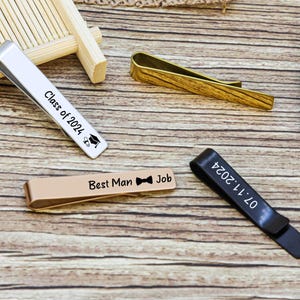 May include: Four metal tie clips in silver, gold, rose gold, and black finishes. The silver clip is engraved with Class of 2024 and a graduation cap. The rose gold clip features Best Man Job with a bow tie icon. The black clip displays 07.11.2024.