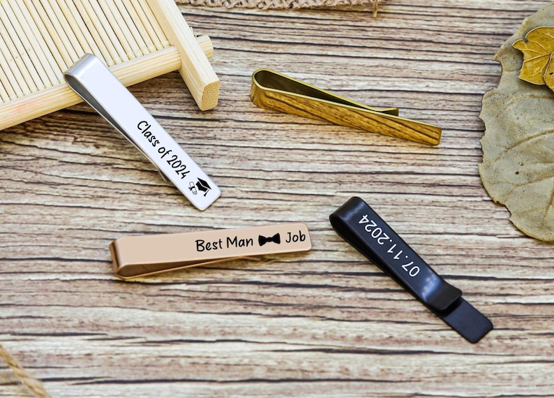 May include: Four metal tie clips in silver, gold, rose gold, and black finishes. The silver clip is engraved with Class of 2024 and a graduation cap. The rose gold clip features Best Man Job with a bow tie icon. The black clip displays 07.11.2024.
