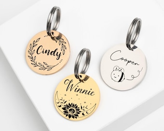 May include: Three gold and silver pet ID tags with key rings. The tags are round and have different designs. One tag has a floral wreath with the name "Cindy" inside. Another tag has a bee with the name "Cooper" and a tail. The third tag has a crescent moon with a sunflower and the name "Winnie".
