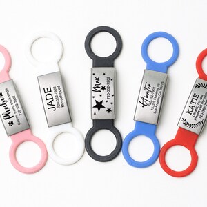 K&ouml;nnte beinhalten: F&uuml;nf personalisierte Haustier-ID-Tags mit verschiedenen Namen und Telefonnummern. Die Tags sind an bunten Silikonringen befestigt.
