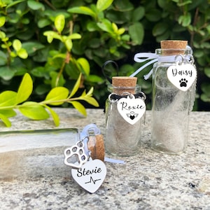 May include: Three small glass jars with cork tops, each containing white fur and a silver charm. The charms are heart-shaped with the names "Stevie", "Roxie", and "Daisy" engraved on them. Each charm also has a small paw print or angel wing design.