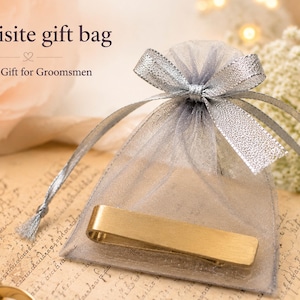 May include: A gold-toned metal tie bar placed in front of a sheer grey organza drawstring gift bag with a silver ribbon bow. The text on the image reads Exquisite gift bag and Perfect Gift for Groomsmen in a dark brown serif font.