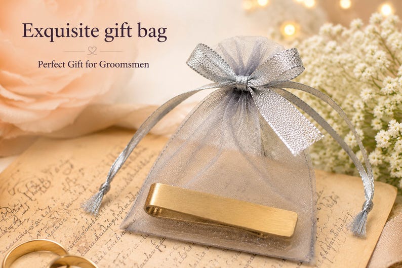 May include: A gold-toned metal tie bar placed in front of a sheer grey organza drawstring gift bag with a silver ribbon bow. The text on the image reads Exquisite gift bag and Perfect Gift for Groomsmen in a dark brown serif font.