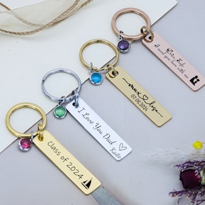 May include: Four gold, silver and rose gold keychains with different engraved messages. The keychains feature a small charm with a coloured gemstone. The messages include "Drive Safe I need you here with me", "max & lisa 07.08.2021", "I Love You Dad Katie", and "Class of 2024".
