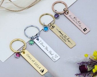 May include: Four gold, silver, and rose gold keychains with different engraved messages. The keychains feature a small charm with a colored gemstone. The messages include "Drive Safe I need you here with me", "max & lisa 07.08.2021", "I Love You Dad Katie", and "Class of 2024".