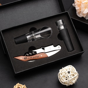 Könnte beinhalten: Eine schwarze Geschenkbox mit schwarzem Schaumstoff-Innenleben, die einen Flaschenöffner, einen Flaschenverschluss und einen Ausgießer enthält. Der Flaschenöffner hat einen Holzgriff mit der Gravur "Grand-pa". Der Flaschenverschluss ist schwarz und der Ausgießer ist aus klarem Glas mit schwarzem Oberteil.