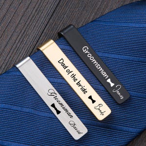 May include: Three metal tie clips in silver, gold, and black finishes, each engraved with text and a bow tie icon. The silver clip reads Groomsman David, the gold clip reads Dad of the bride Bride, and the black clip reads Groomsman James.