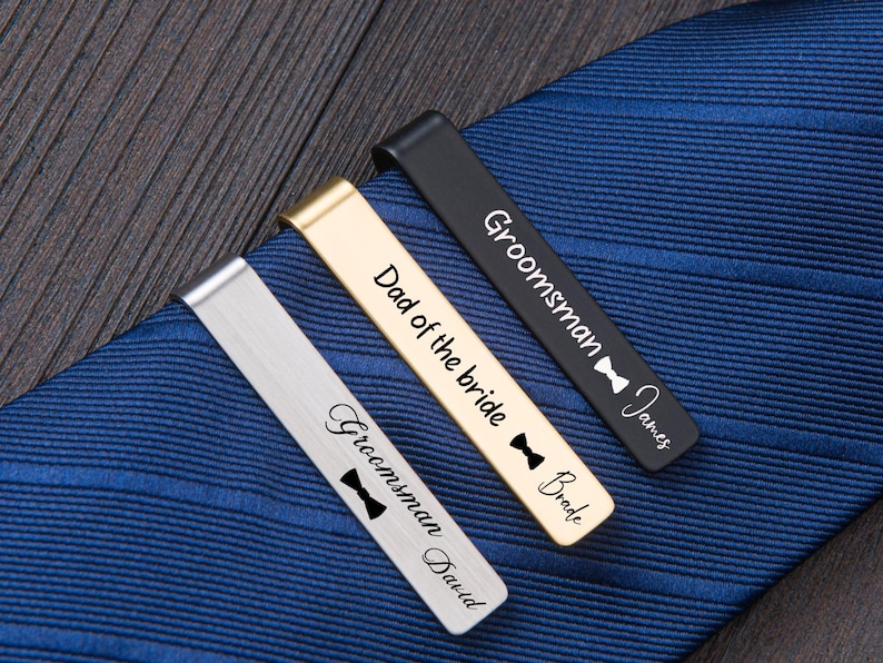 May include: Three metal tie clips in silver, gold, and black finishes, each engraved with text and a bow tie icon. The silver clip reads Groomsman David, the gold clip reads Dad of the bride Bride, and the black clip reads Groomsman James.