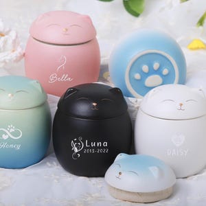 Peut inclure: Cinq urnes en céramique en forme de chat avec couvercles, chacune avec une couleur et un design différents. Les urnes sont étiquetées "Bella", "Honey", "Luna 2013-2022", "Daisy" et une empreinte de patte.