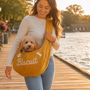 Puede incluir: Un bolso bandolera de color amarillo mostaza para mascotas con la palabra Biscuit en letras cursivas blancas, un aplique de flor blanca y un aplique de hueso blanco. El bolso de tela tiene una correa ancha y contiene un perro pequeño de pelo rizado.