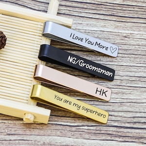 May include: Four metal tie clips in silver, black, rose gold, and gold finishes. The silver clip is engraved with "I Love You More" and a heart, the black with "NG/Groomsman", the rose gold with "HK", and the gold with "You are my superhero".