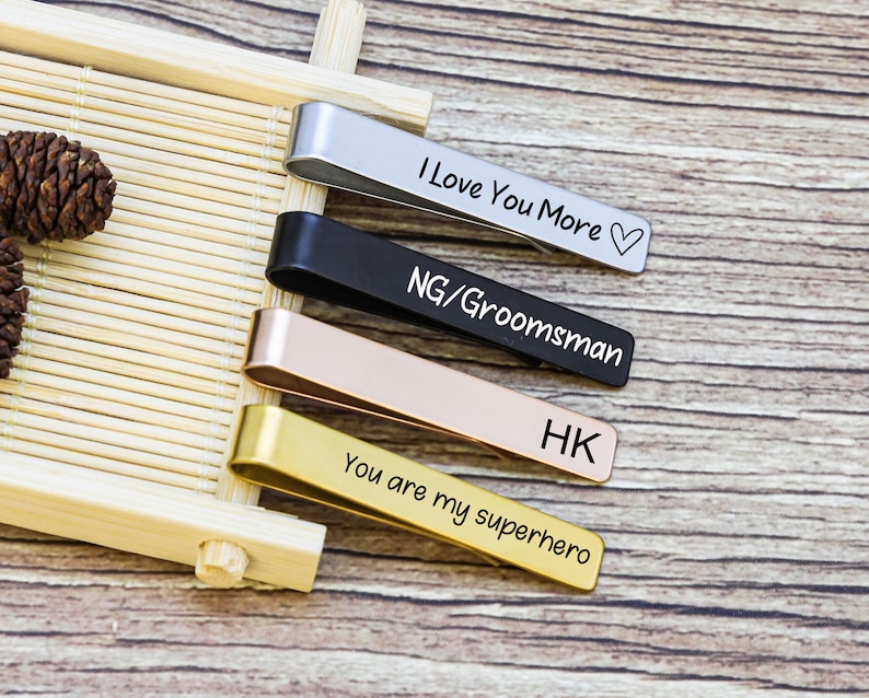 May include: Four metal tie clips in silver, black, rose gold, and gold finishes. The silver clip is engraved with "I Love You More" and a heart, the black with "NG/Groomsman", the rose gold with "HK", and the gold with "You are my superhero".