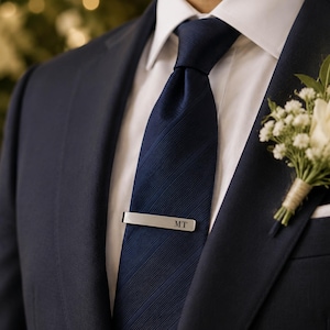 May include: A navy blue suit jacket worn over a crisp white collared shirt and a dark blue diagonal-striped necktie. A silver-toned metal tie bar engraved with the letters MT is clipped to the tie. A white rose boutonniere is pinned to the lapel.