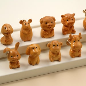 Peut inclure: Une collection de onze petites figurines d'animaux en bois. Les figurines comprennent un serpent, une souris, un cochon, un tigre, un poulet, un lapin, un chien, un singe, une vache et un mouton. Chaque figurine est de couleur marron clair et possède une petite vis métallique.