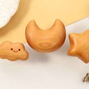 May include: Wooden cloud, crescent moon, and star-shaped toys. The cloud has a smiling face. The toys are made of light-coloured wood and are arranged on a white and yellow background. A decorative plate and pinecone are also visible.