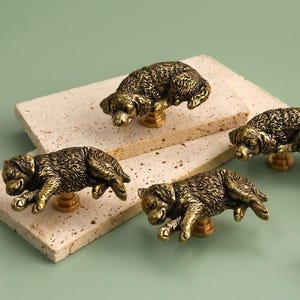 May include: Four brass-toned metal cabinet knobs, each shaped like a small dog lying down. The figures feature detailed fur textures and a metallic finish. Each knob is attached to a circular base, designed for mounting onto furniture or cabinetry.