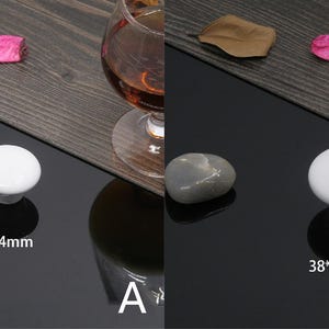 K&ouml;nnte beinhalten: Zwei wei&szlig;e Keramik-Schrankkn&ouml;pfe, einer mit den Ma&szlig;en 32 mm x 24 mm und der andere mit 38 mm x 28 mm, werden auf einer gl&auml;nzenden schwarzen Oberfl&auml;che pr&auml;sentiert. Ein grauer Stein und ein Glas bernsteinfarbener Fl&uuml;ssigkeit sind ebenfalls zu sehen.