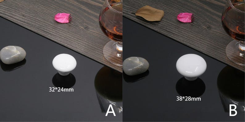 K&ouml;nnte beinhalten: Zwei wei&szlig;e Keramik-Schrankkn&ouml;pfe, einer mit den Ma&szlig;en 32 mm x 24 mm und der andere mit 38 mm x 28 mm, werden auf einer gl&auml;nzenden schwarzen Oberfl&auml;che pr&auml;sentiert. Ein grauer Stein und ein Glas bernsteinfarbener Fl&uuml;ssigkeit sind ebenfalls zu sehen.