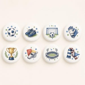 Pode incluir: Um conjunto de oito botões redondos brancos com ilustrações de futebol, incluindo uma bola, chuteiras, uma baliza, luvas de guarda-redes, um troféu dourado, um apito com cartões vermelho e amarelo, um estádio e um cachecol às riscas com uma bandeira.