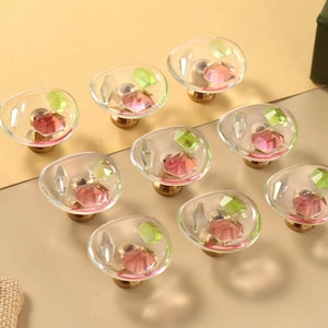 Può includere: Un set di nove pomelli rotondi per mobili con parte superiore in vetro trasparente e cristalli sfaccettati rosa e verde chiaro incastonati. Ogni pomello presenta una base in metallo dorato e una forma circolare leggermente irregolare.
