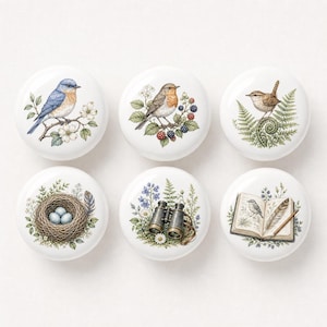 May include: A set of six round white ceramic knobs featuring detailed illustrations. Designs include a bluebird on a branch, a robin with blackberries, a wren on a fern, a bird nest with eggs, binoculars with wildflowers, and an open book with a quill.